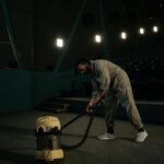 A cinema employee cleaning the theater floor with a vacuum cleaner under dim lights.